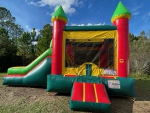 An inflatable bounce house and slide combo featuring bright green, yellow and red colors. The structure includes a large bounce area, a climbing wall, and a slide, with netted windows for visibility and airflow. Set up on a grassy lawn with trees in the background, the scene is lively and inviting, ideal for outdoor fun.