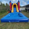 A vibrant inflatable bounce house and slide combo set up outdoors on a grassy lawn. The structure showcases bold primary colors of red, blue, and yellow, with a spacious bouncing area, a climbing wall, and a slide. The bounce house has mesh windows for ventilation and visibility, while the surrounding green grass and clear sky enhance the cheerful and fun atmosphere of the scene.