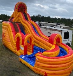 A dramatic, climbing, inflatable water slide called 'Fire Island,' showcasing vibrant colors of orange, yellow, and blue. The slide features a steep, curving descent that ends in a splash pool. Set up on a grassy lawn.