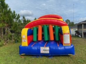 A large inflatable obstacle course set up outdoors, featuring vibrant colors of blue, green, yellow, and red. The course includes various challenges such as tunnels, climbing walls, and slides, designed for active play. The structure is laid out on a grassy area, with trees in the background, creating an exciting and adventurous environment for participants."