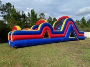 Side view of a large inflatable obstacle course set up outdoors, featuring vibrant colors of blue, green, yellow, and red. The course includes various challenges such as tunnels, climbing walls, and slides, designed for active play. The structure is laid out on a grassy area, with trees in the background, creating an exciting and adventurous environment for participants."