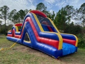 A large inflatable obstacle course set up outdoors, featuring vibrant colors of blue, green, yellow, and red. The course includes various challenges such as tunnels, climbing walls, and slides, designed for active play. The structure is laid out on a grassy area, with trees in the background, creating an exciting and adventurous environment for participants."